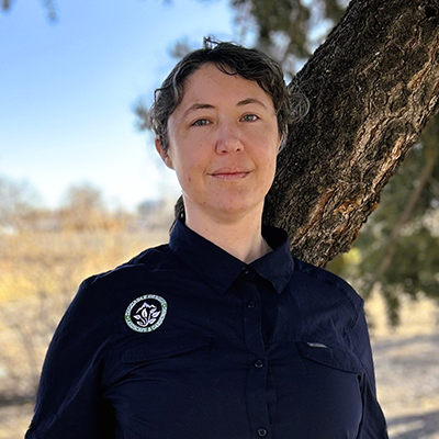 Woman in navy shirt with logo, leaning against a tree outdoors.