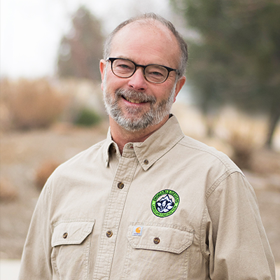 Man wearing glasses and tan shirt with logo outdoors smiling.
