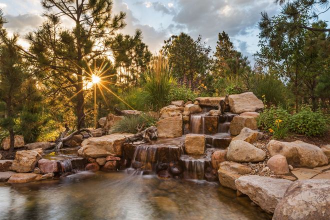A sunlit rock waterfall flows into a pond, surrounded by lush trees and greenery at golden hour.