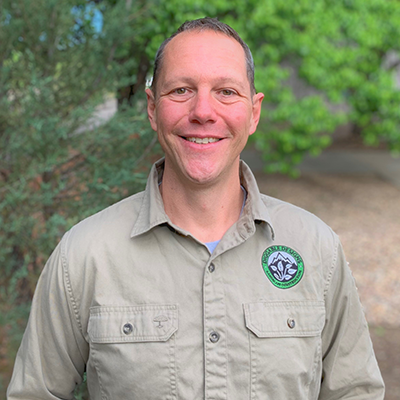 Man in khaki shirt with logo smiles outdoors, blurry green background.