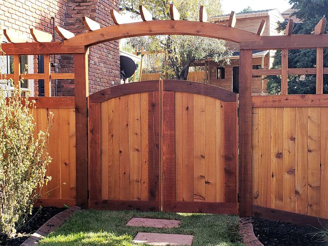 A natural wood double gate featuring an arched pergola top, set into a matching cedar fence in a residential garden.