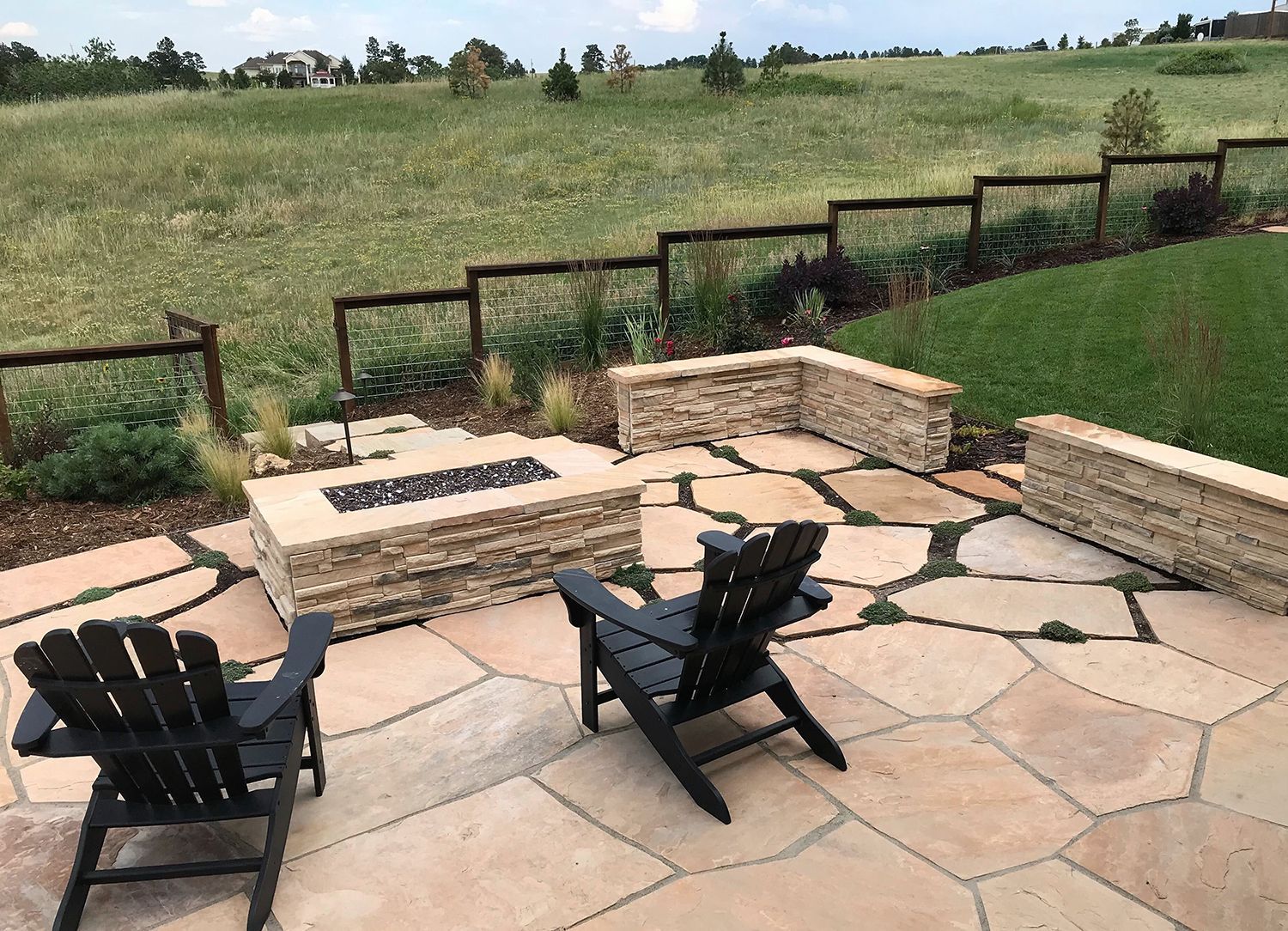 Two black Adirondack chairs face a stone fire pit on a flagstone patio overlooking a grassy field with a wood fence.