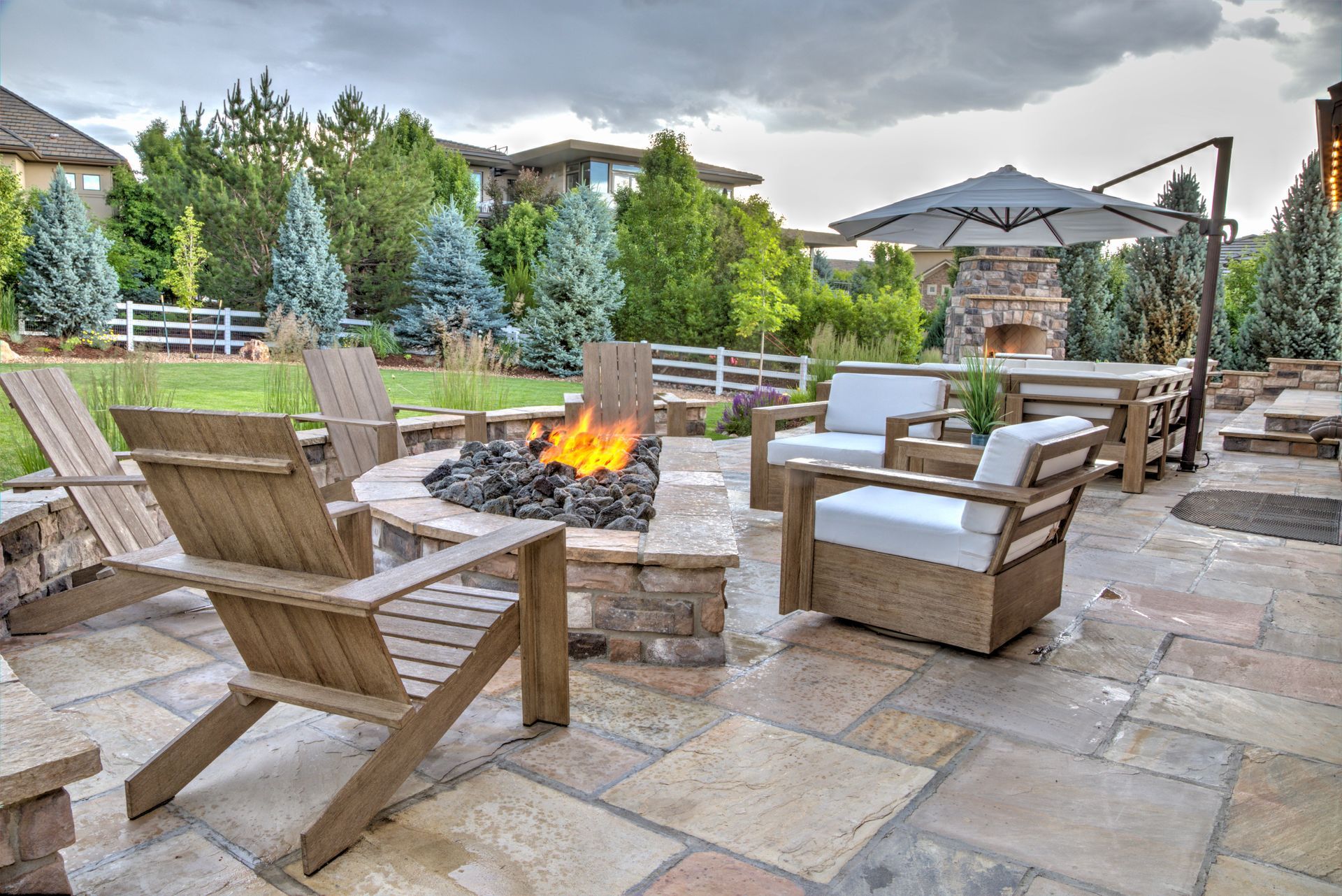 Patio with fire pit, seating, table, umbrella; stone pavers, green grass, trees, and cloudy sky.
