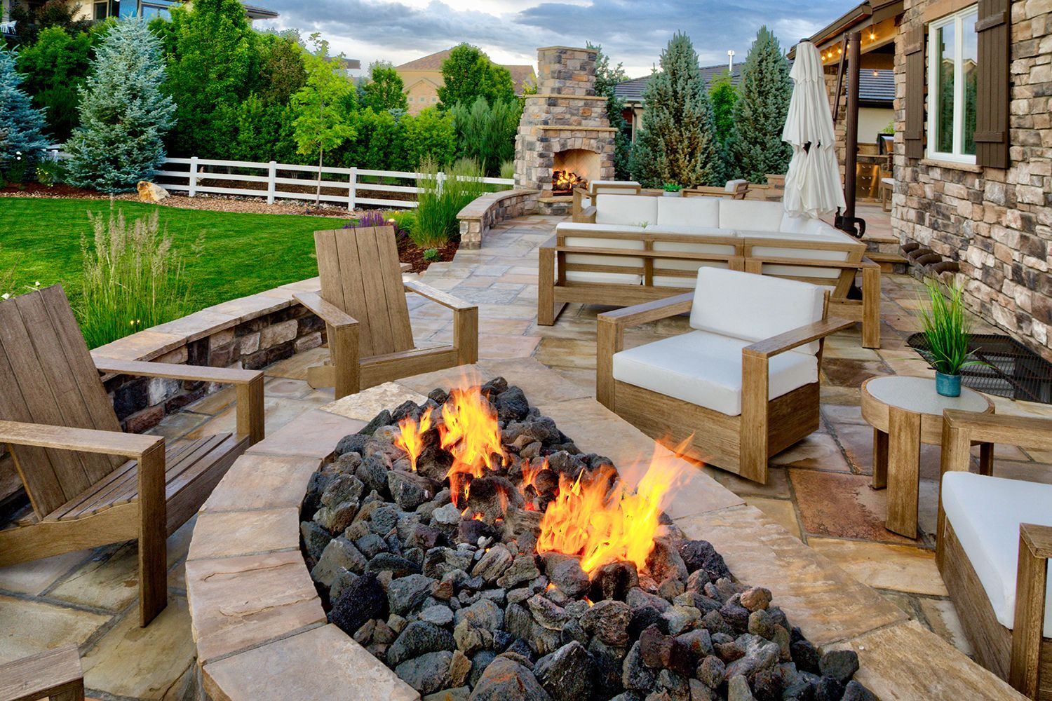 A backyard stone patio featuring a lit fire pit, outdoor lounge furniture, and a stone fireplace at dusk.