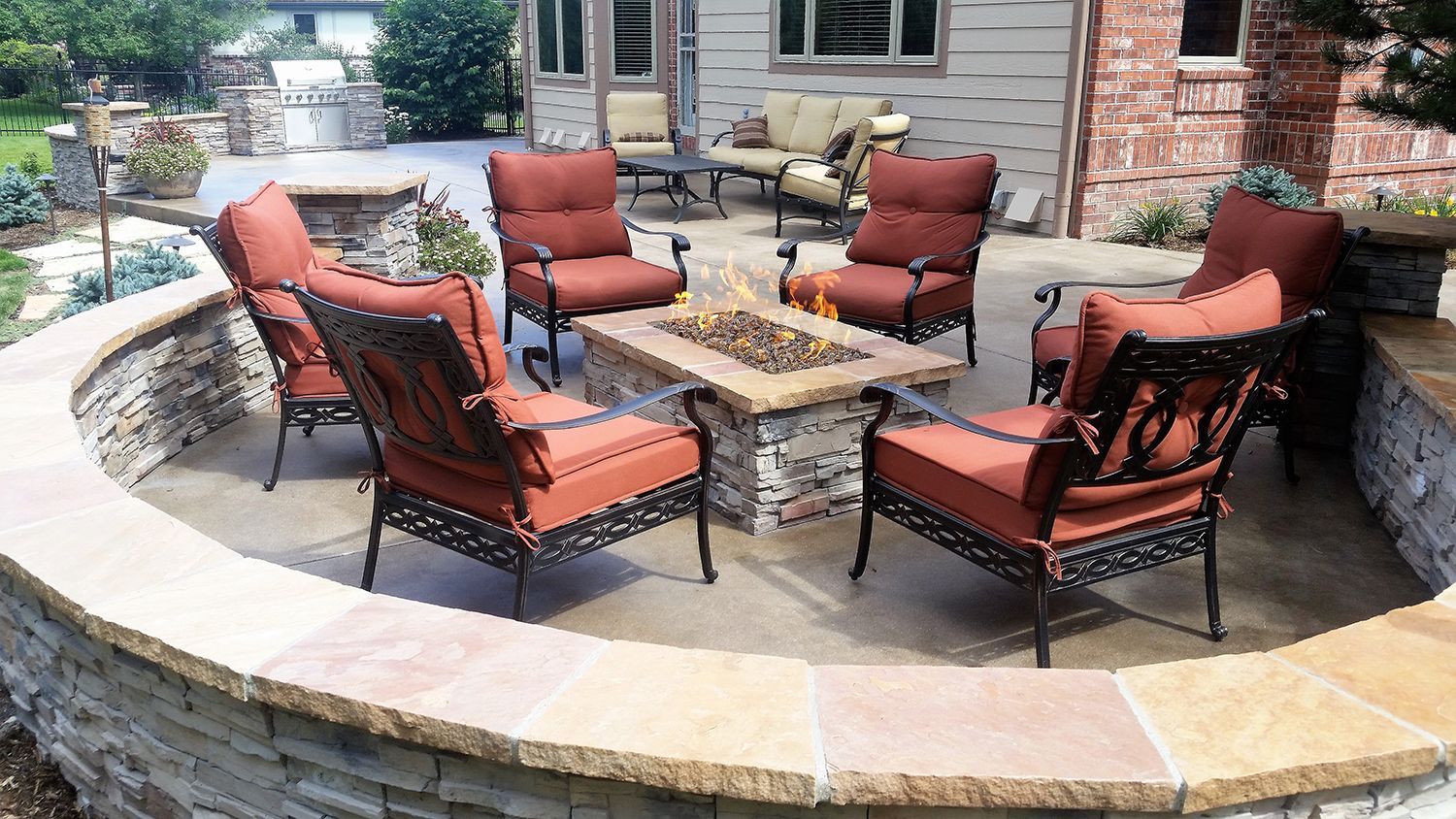 A stone patio with a central fire pit surrounded by six matching metal-framed chairs with orange cushions.