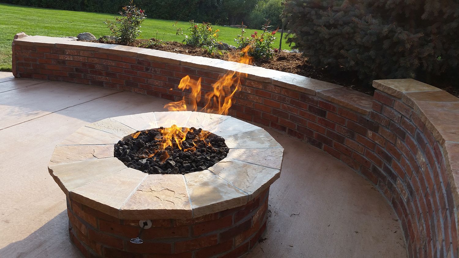 A fire pit with a light stone rim and glowing flames, set on a patio in front of a curved brick retaining wall.