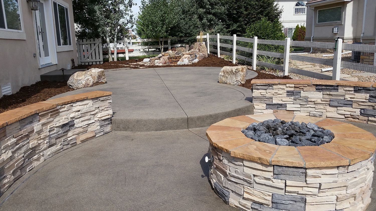 A backyard patio with a stone fire pit, curved stone seating walls, and a concrete surface next to a house.