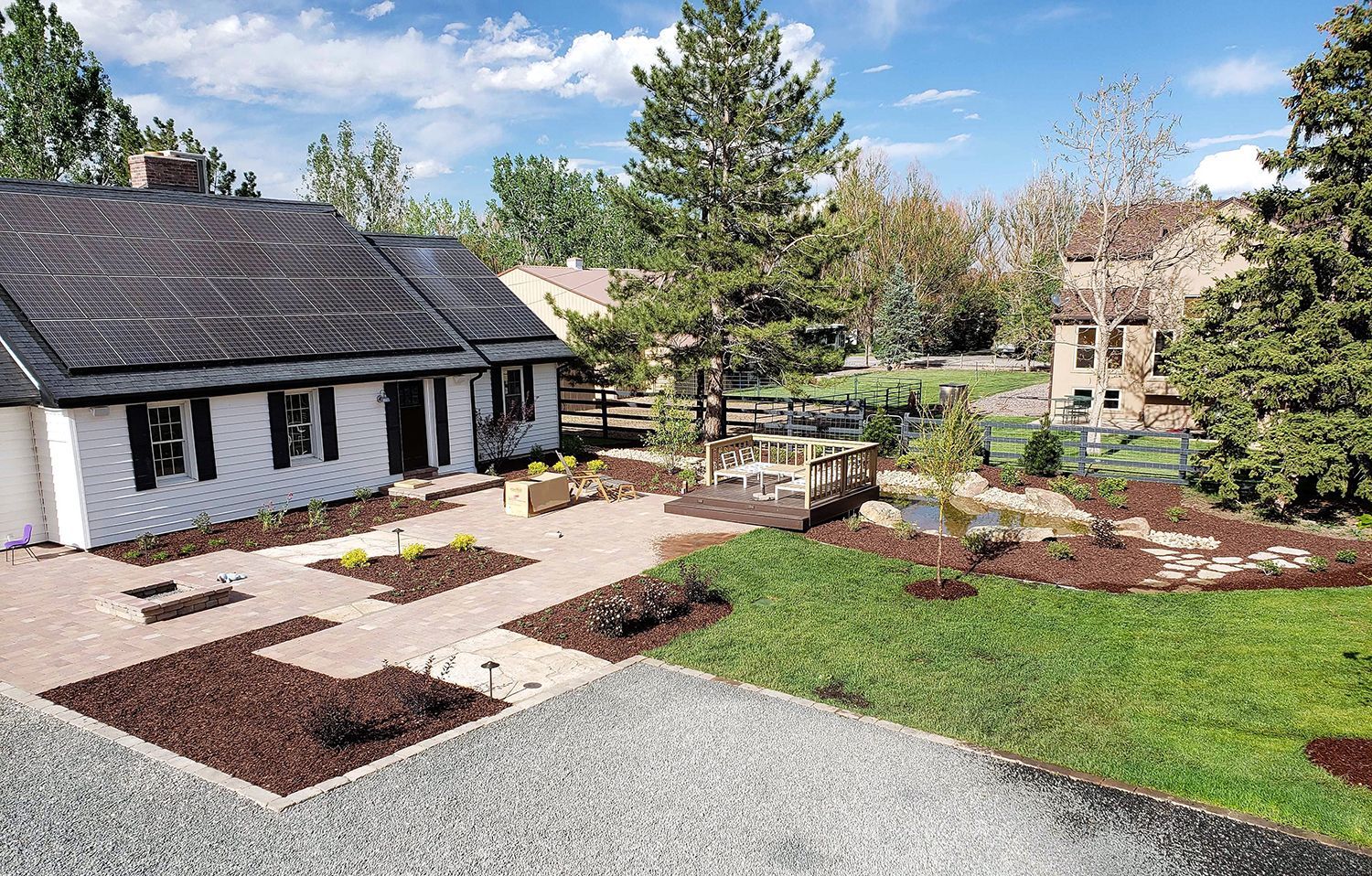 A white cottage with a dark roof stands beside a garden featuring brick paths, mulch beds, and a small wooden bridge.