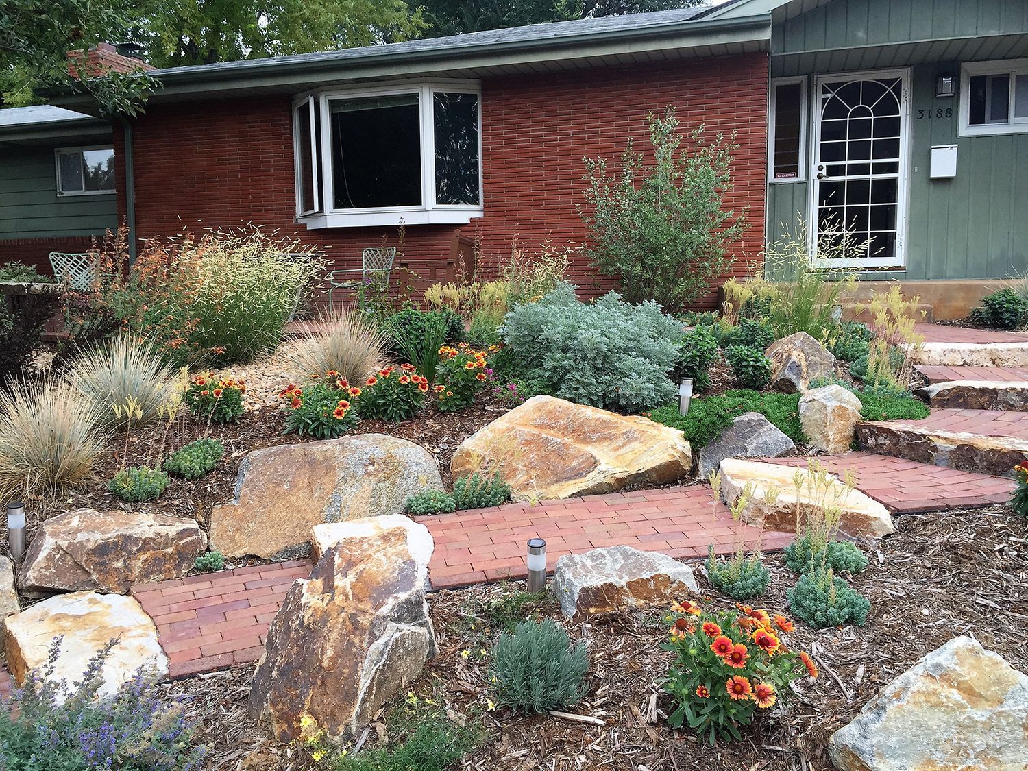 A house with red brick siding, a brown front door, and a landscaped yard with large boulders and orange flowers.