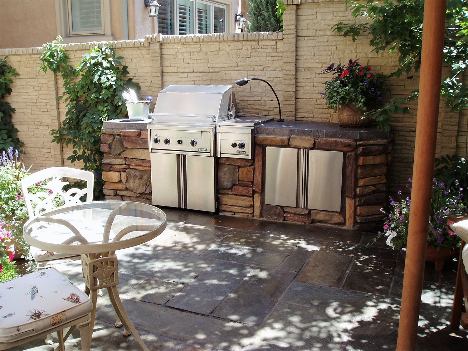 Outdoor kitchen with a built-in stainless steel grill, stone counters, and a small bistro table on a paved patio.