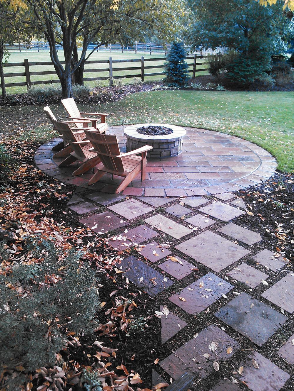 A circular stone fire pit patio with two wooden Adirondack chairs, set in a backyard with a stone path and wooden fence.