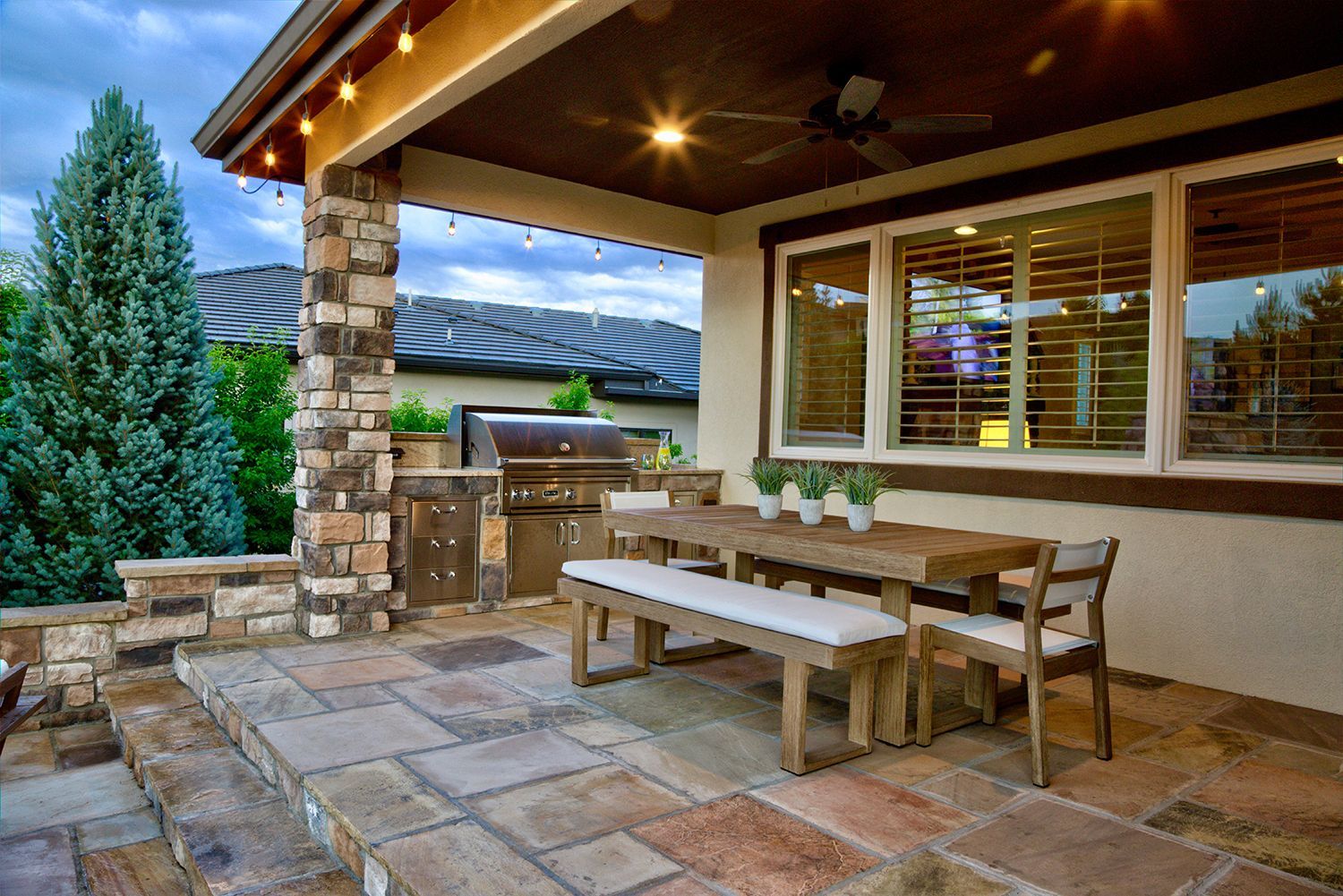 Covered patio with a stone dining set, a built-in grill, and string lights against a background of trees and a house.