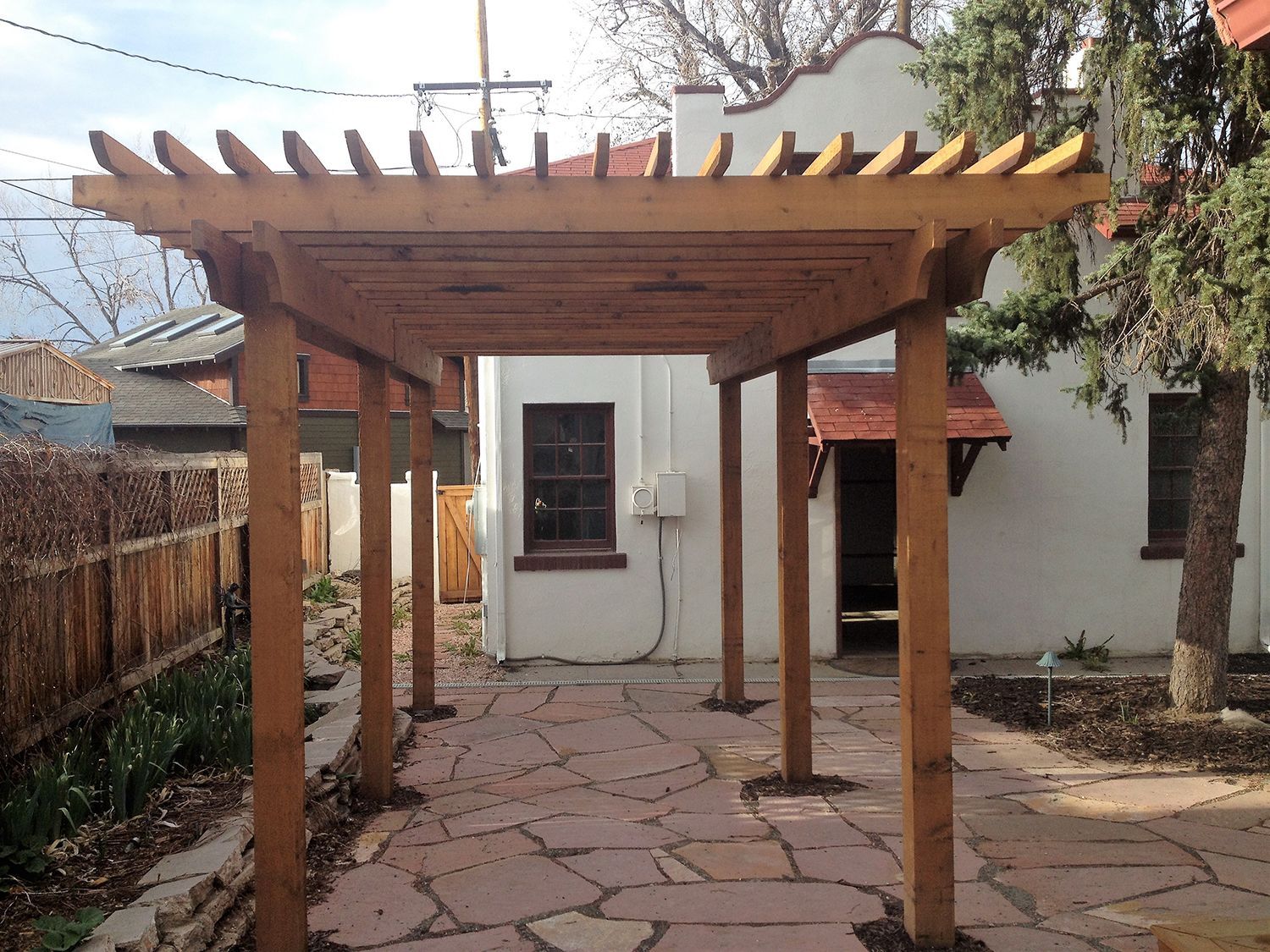 A wooden pergola stands on a flagstone patio in a backyard beside a white stucco house.