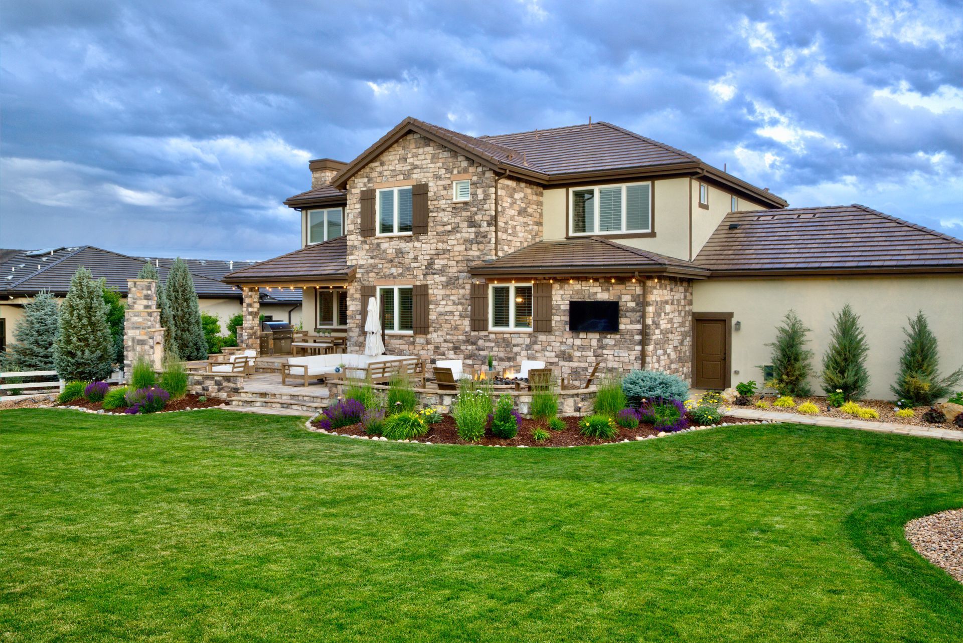A two-story stone house with a patio and outdoor TV sits behind a lush green lawn under a stormy, cloud-filled sky.