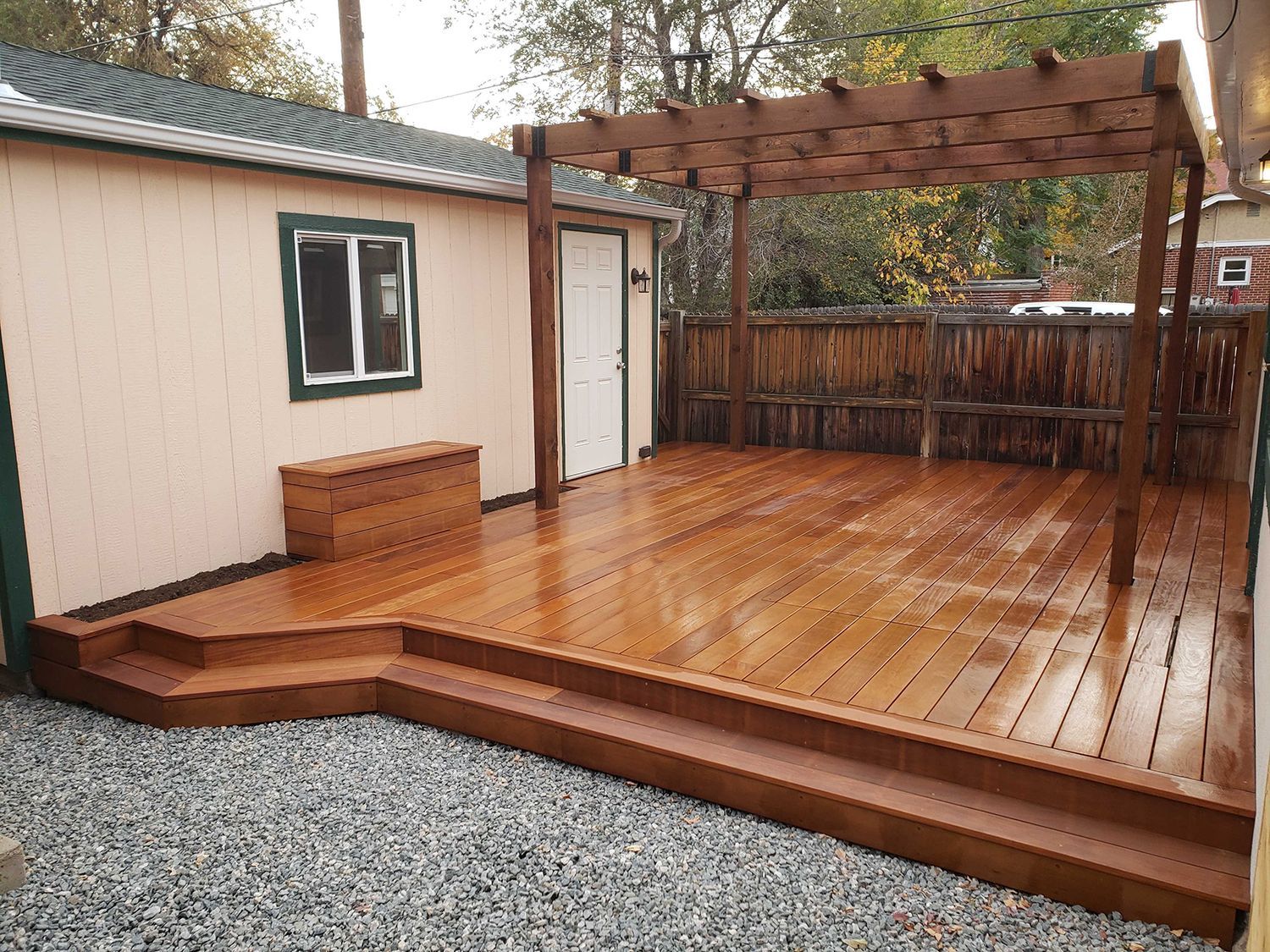 A newly stained wooden deck with a matching pergola and built-in storage box, situated next to a light-colored building.