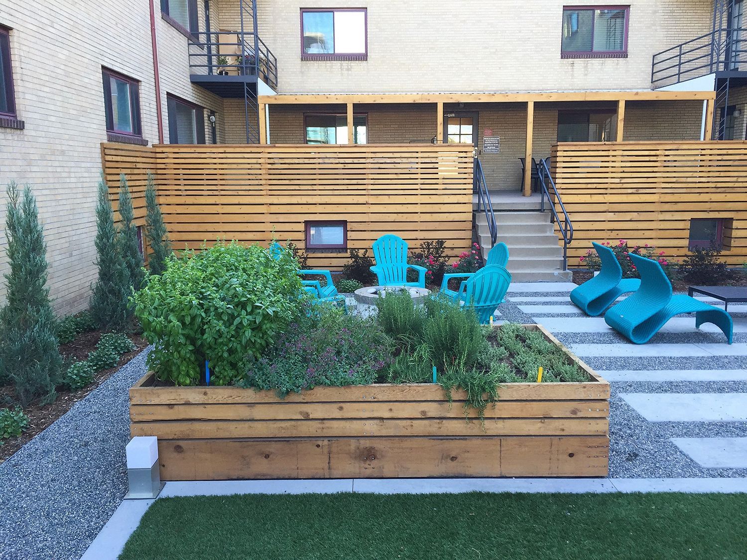 A modern apartment courtyard features a raised wooden garden bed, teal lounge chairs, and a gravel patio.
