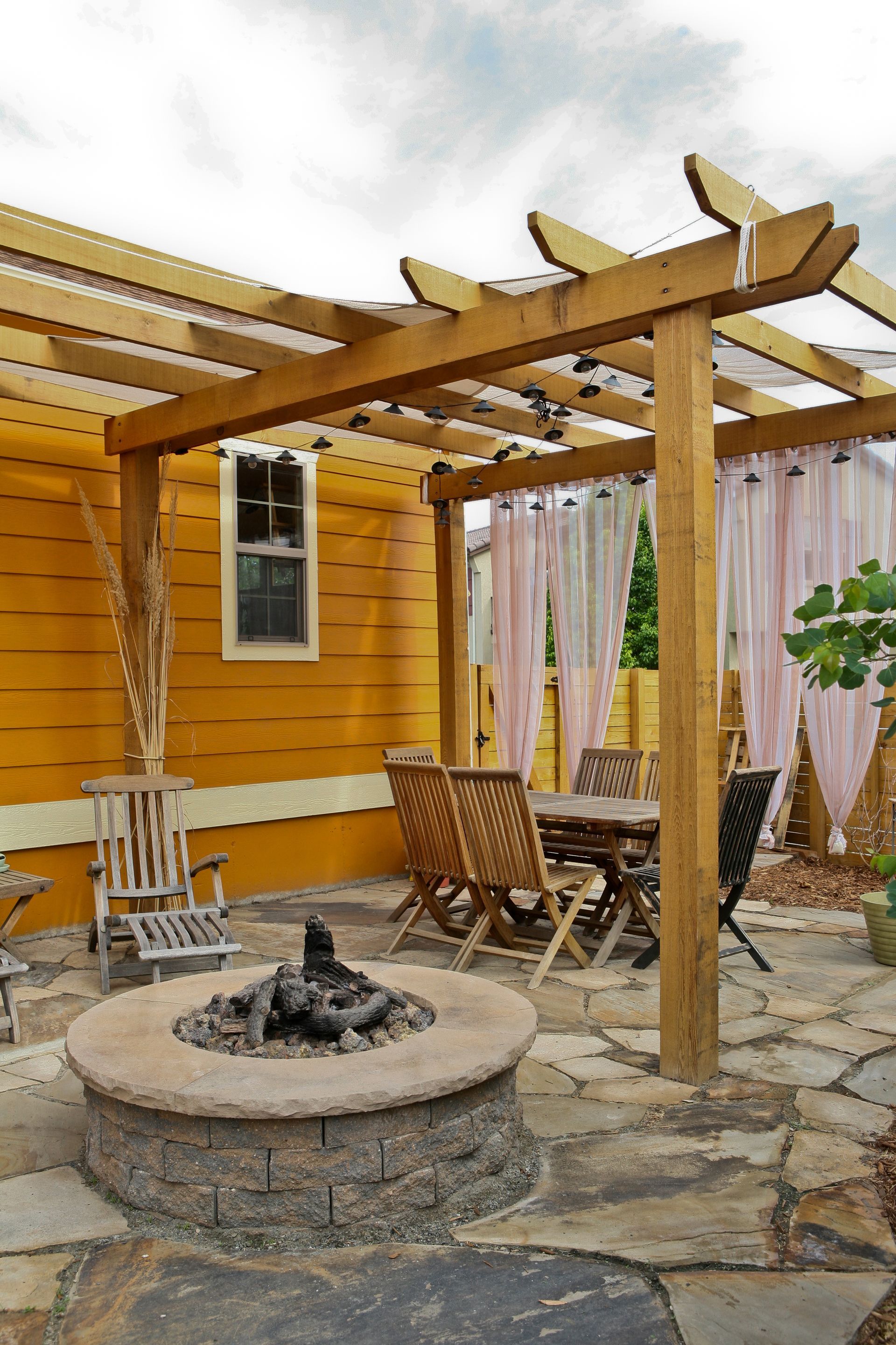 Backyard patio with fire pit, wooden pergola, and seating near a yellow house.