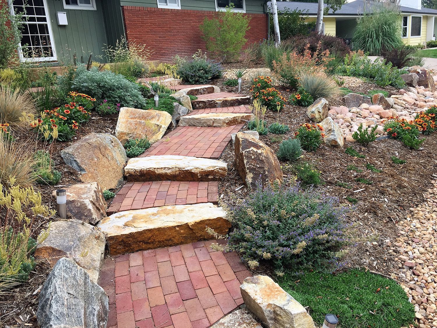 A brick pathway with large stone steps leading up to a green house, surrounded by landscaped garden beds and mulch.
