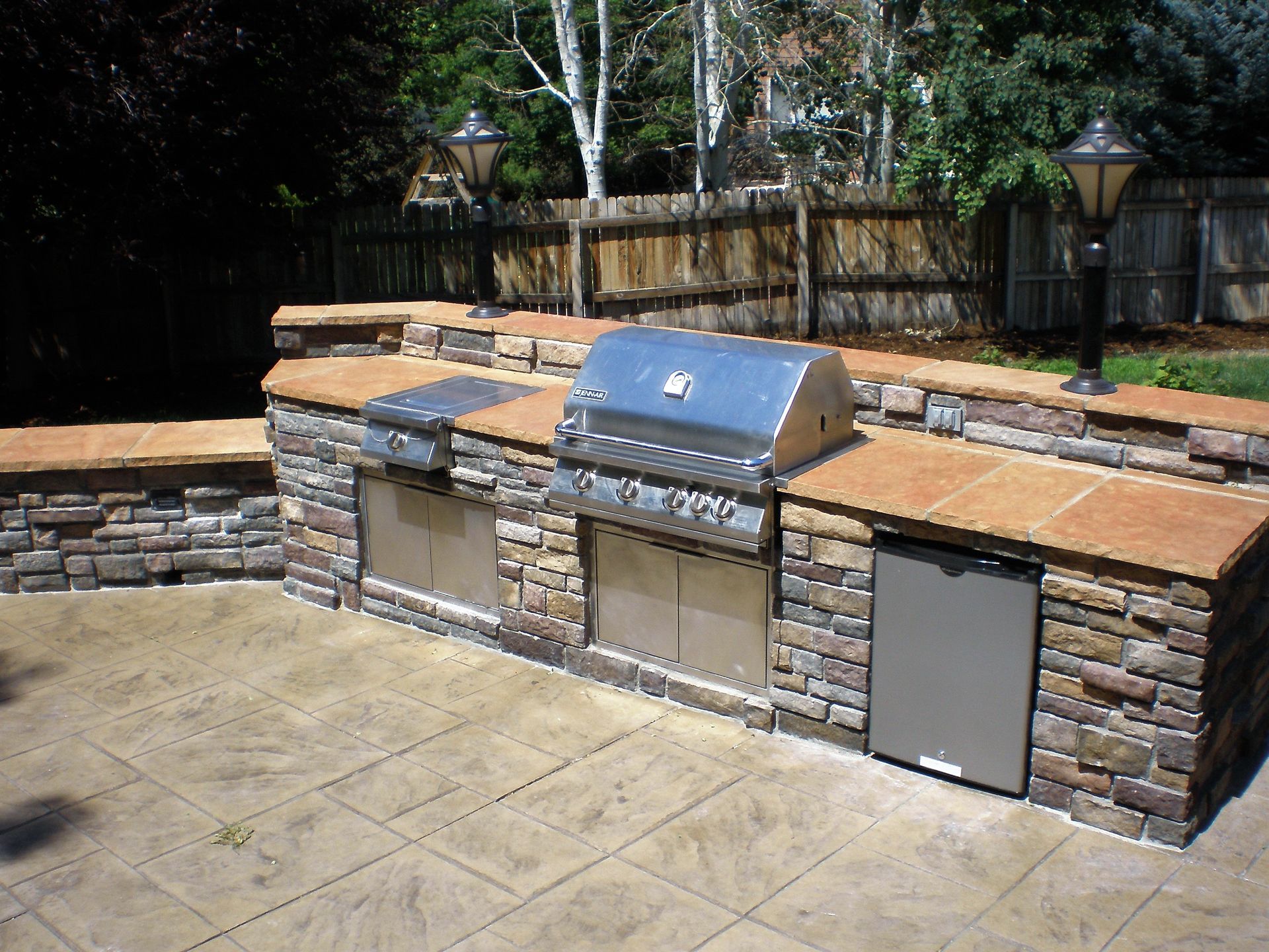 Outdoor stone built-in kitchen with grill, side burner, refrigerator, and counters, in a backyard setting.