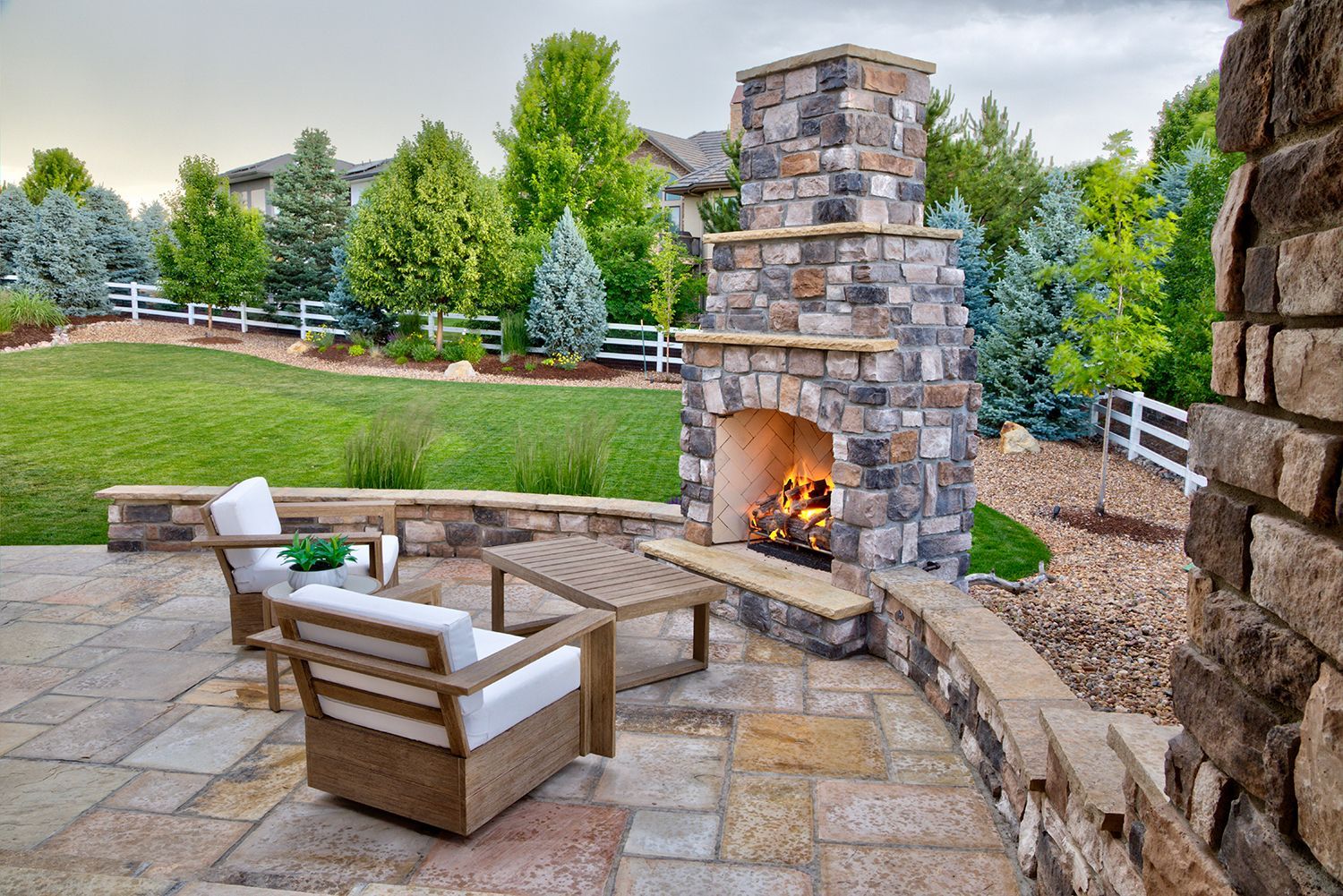 A patio with a lit stone fireplace, two wicker chairs, and a coffee table overlooking a grassy yard with trees and a fence.
