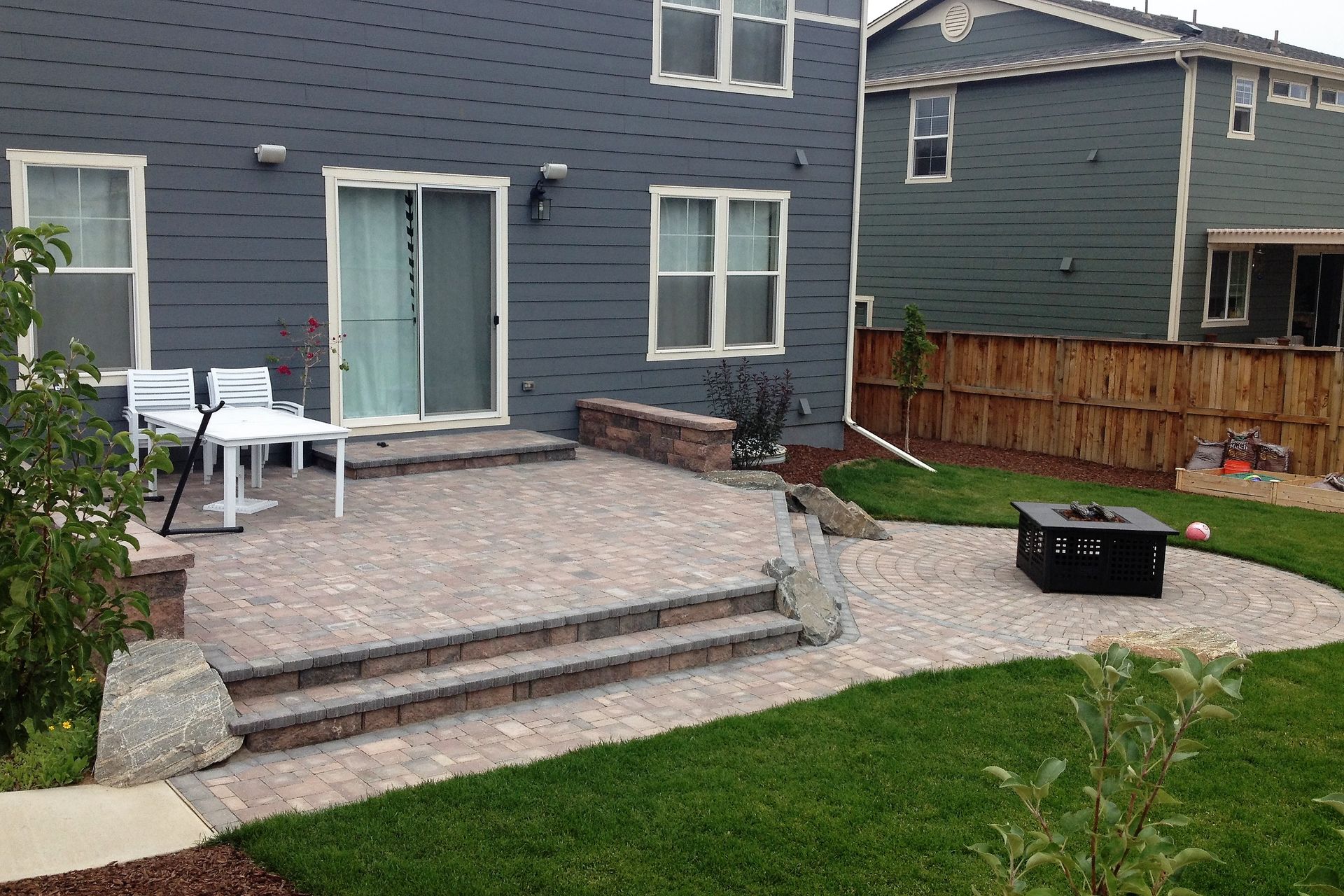 Backyard patio with brick pavers, steps, fire pit, and adjacent grass. House in the background.