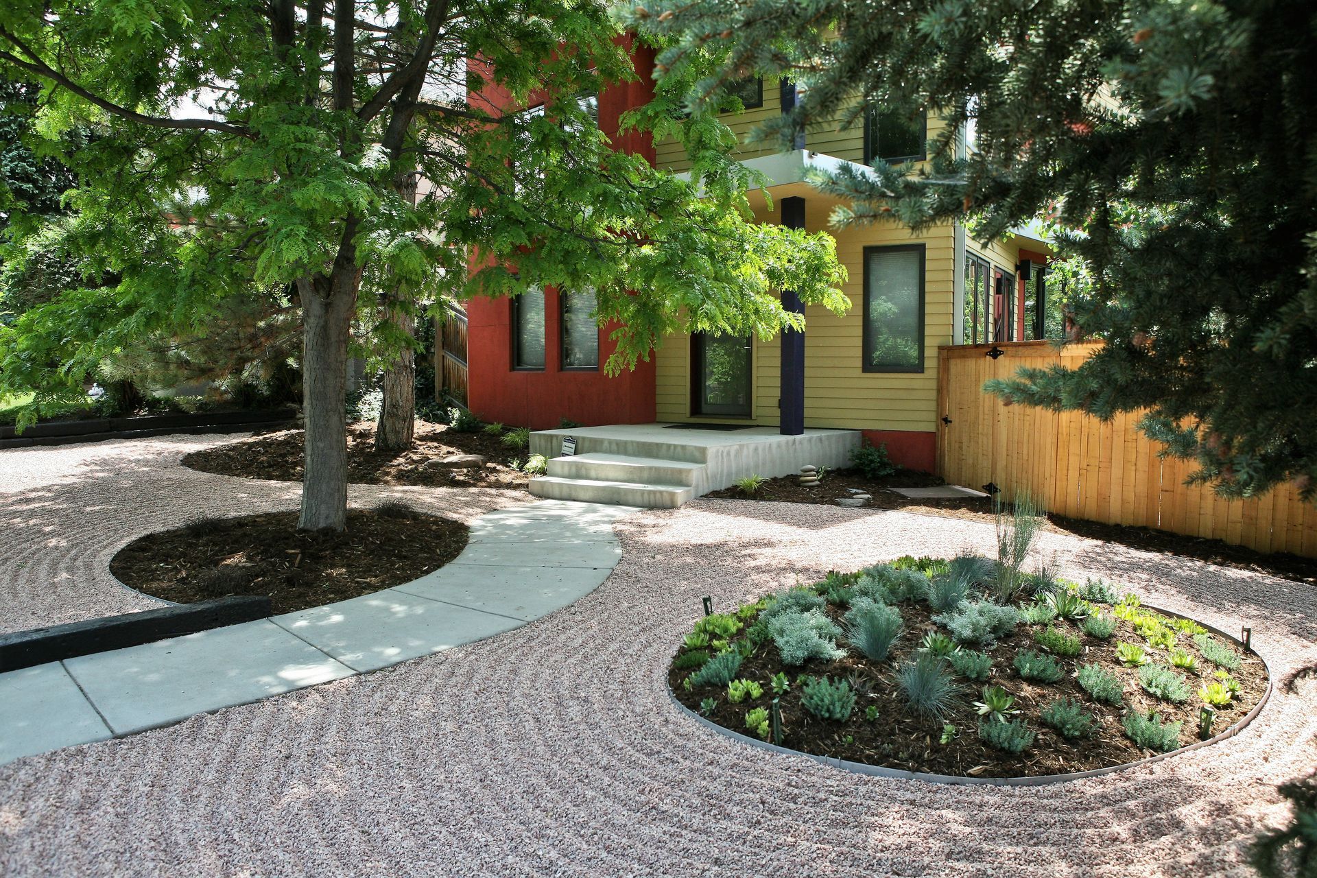 Colorful house with a concrete walkway and gravel yard, trees, and a small garden bed.