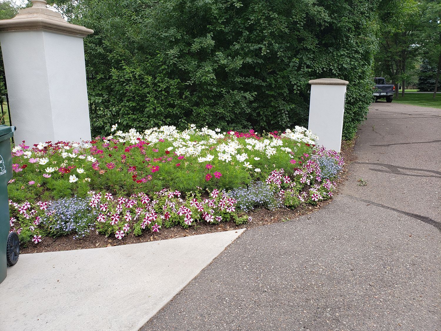 A flower bed with white, pink, and purple flowers situated between two white stone pillars near a paved driveway.