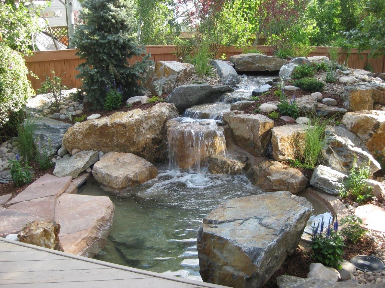 A tiered garden waterfall flowing into a small pond, surrounded by large rocks, greenery, and a stone patio.