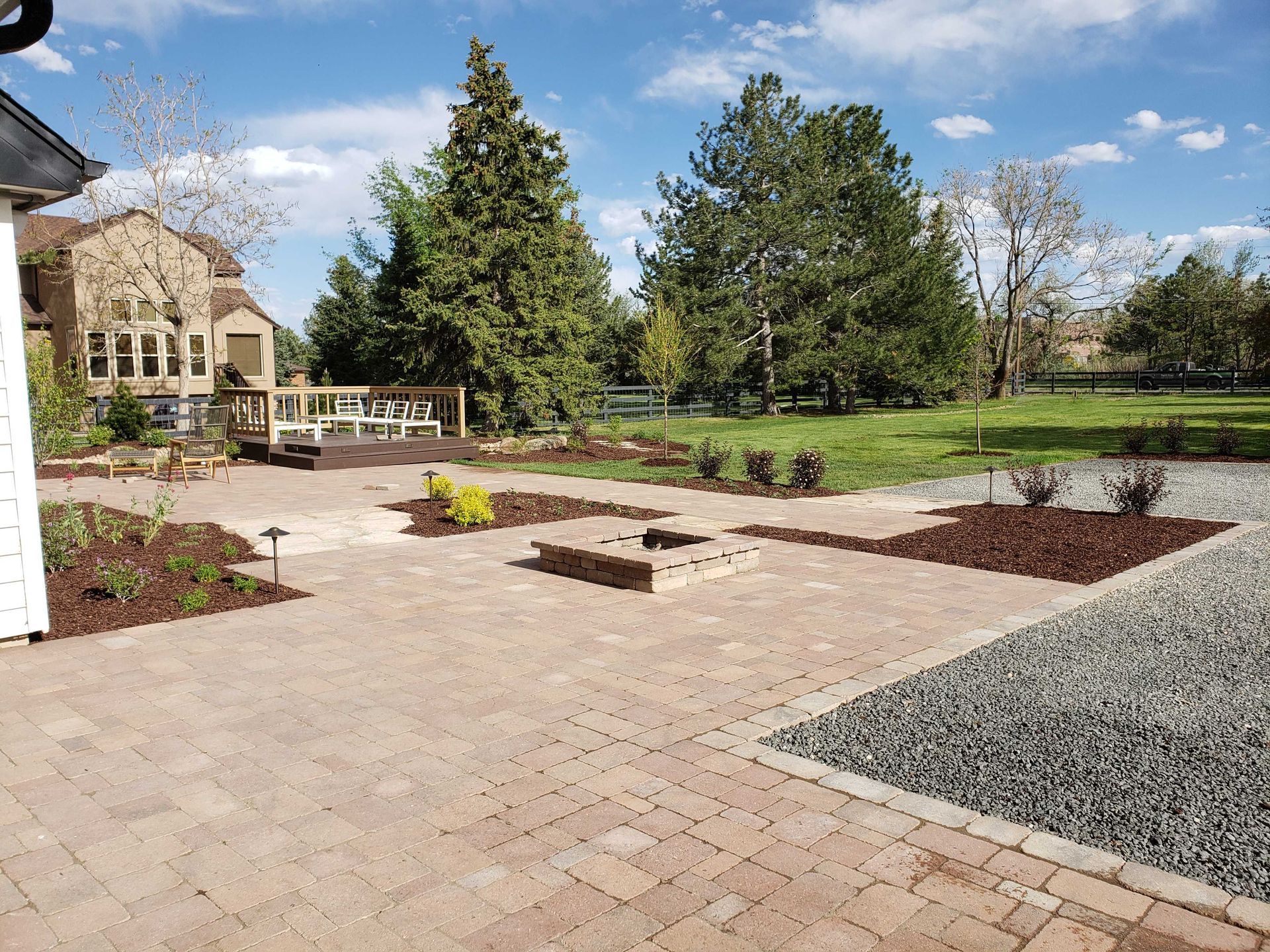 Backyard patio with brick pavers, fire pit, and landscaping, against green lawn and trees.