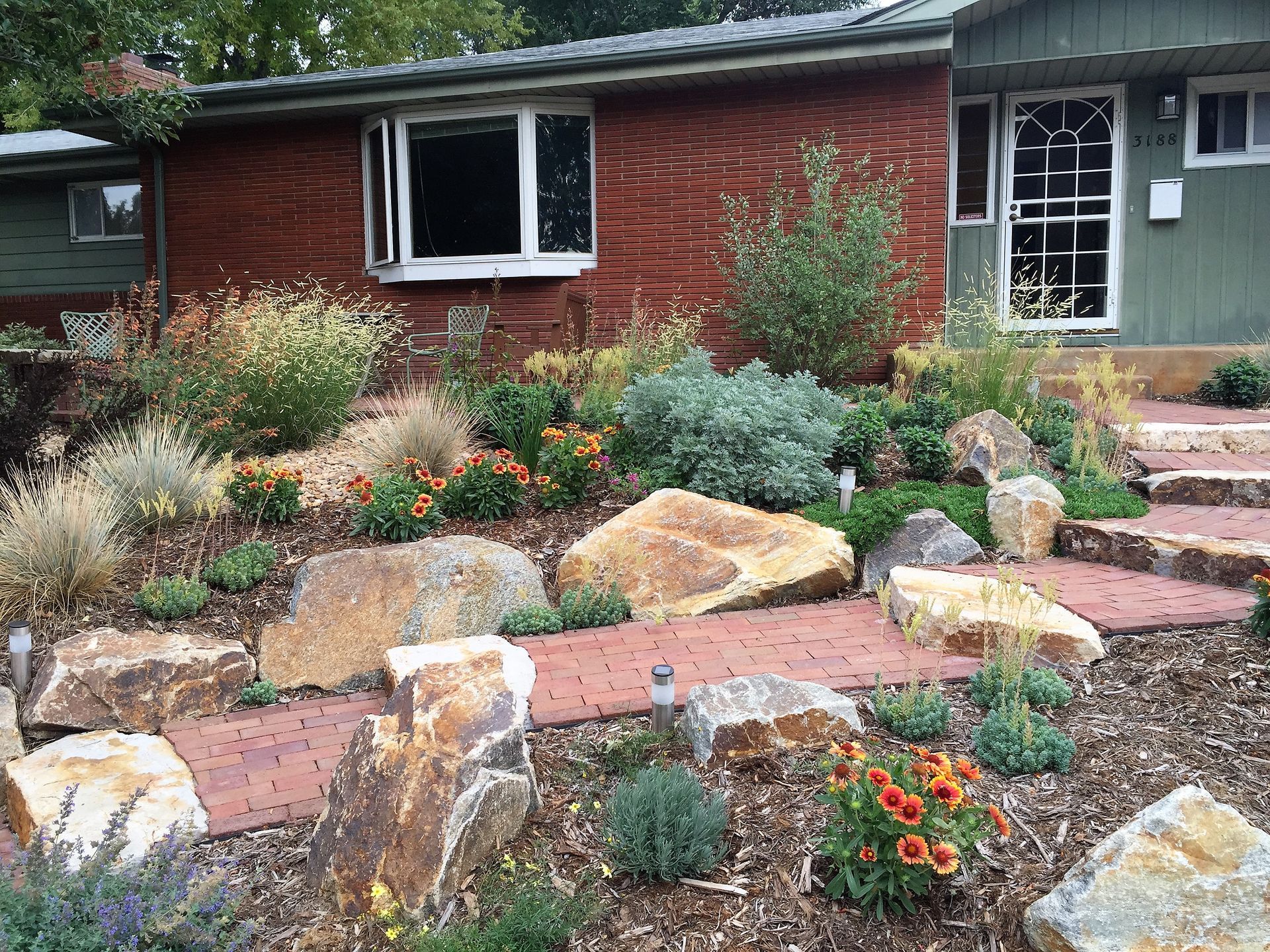 A house with a red-brown exterior, surrounded by a rock garden with various plants and boulders.