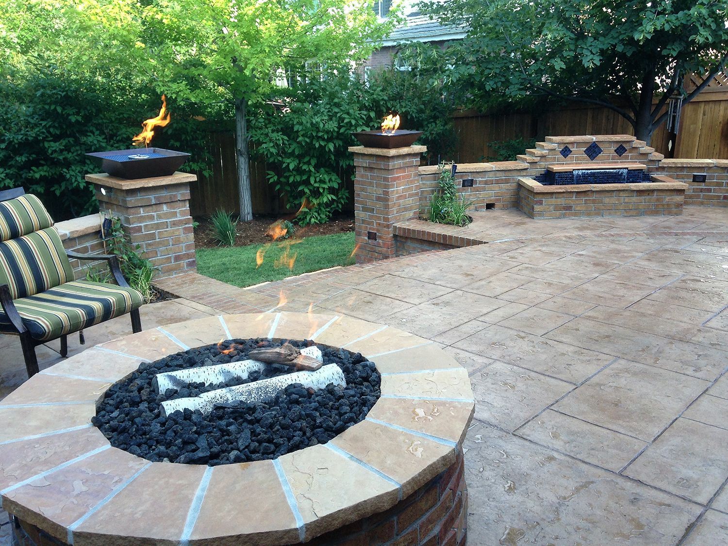 A patio featuring a circular stone fire pit, two brick fire columns with flames, and a stone water fountain in the back.