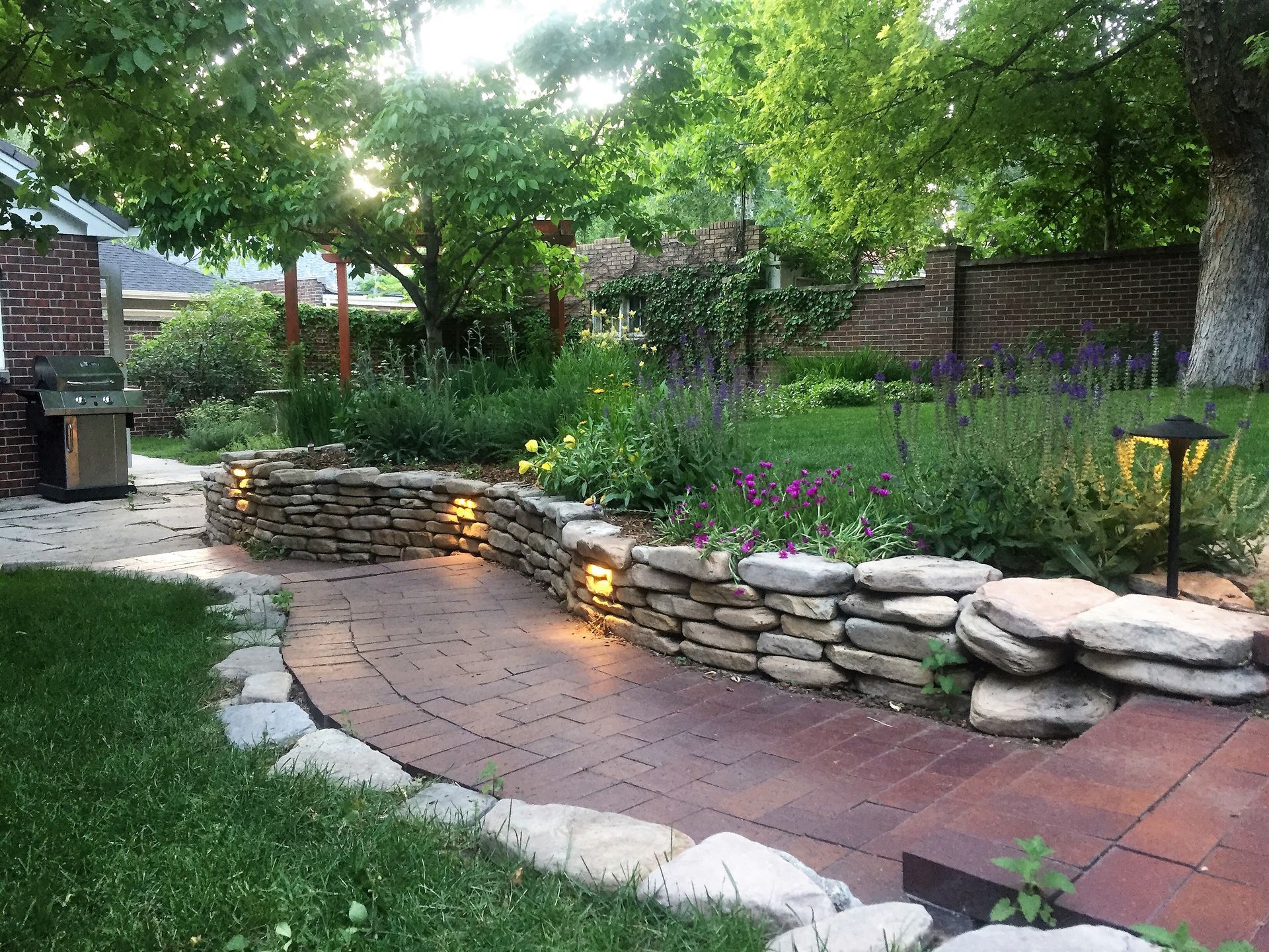 Stone pathway curves through a lush garden with a stone wall and built-in lighting. Green foliage surrounds.
