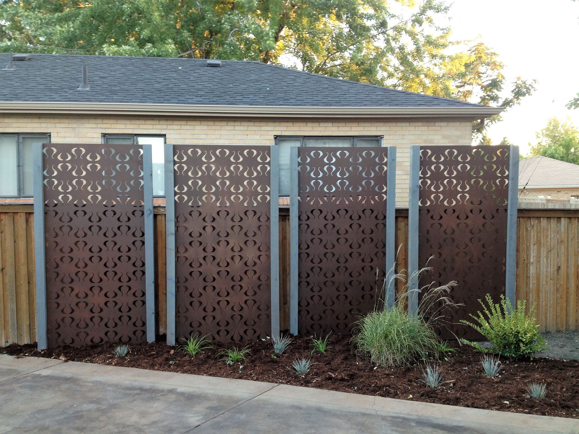 Four brown patterned metal panels on grey supports create a privacy screen in front of a house.