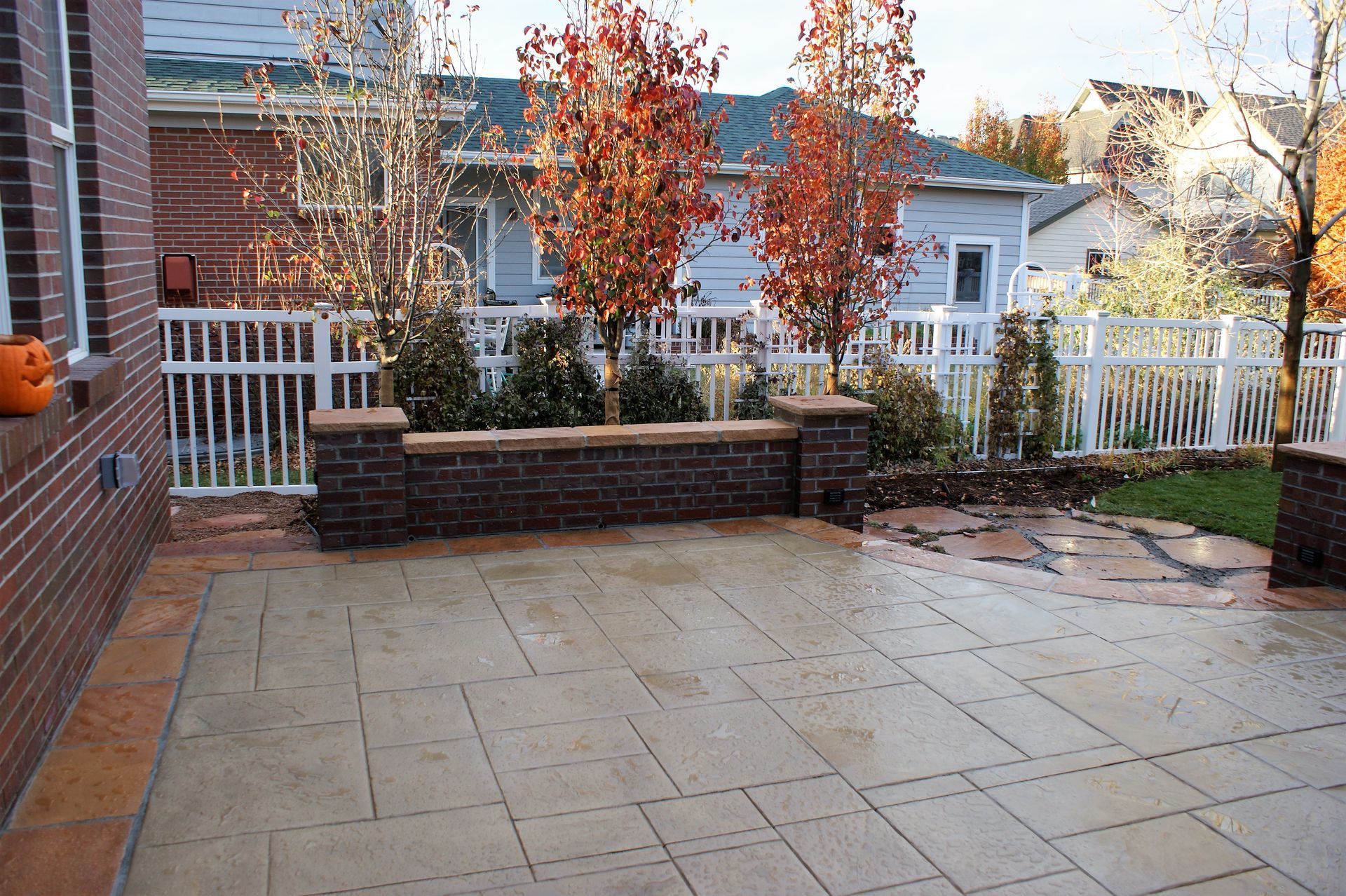 Brick patio with a retaining wall, trees, and white fence in a backyard setting.
