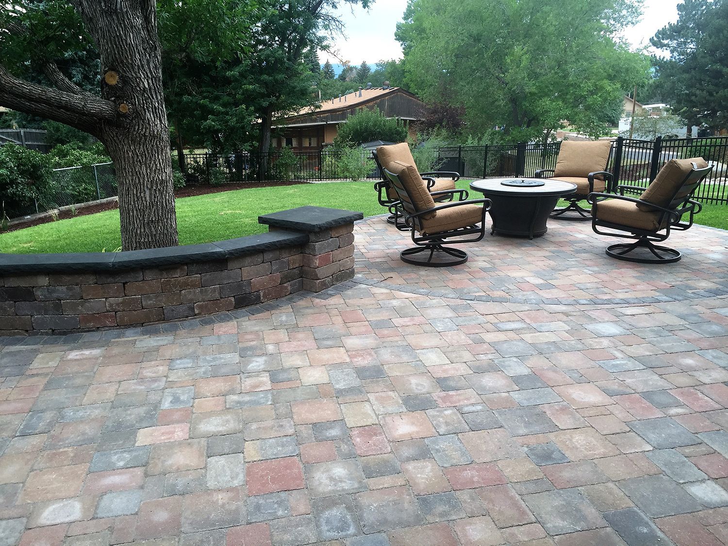 Outdoor patio featuring multicolored brick pavers, a curved brick retaining wall, a fire pit, and four cushioned chairs.