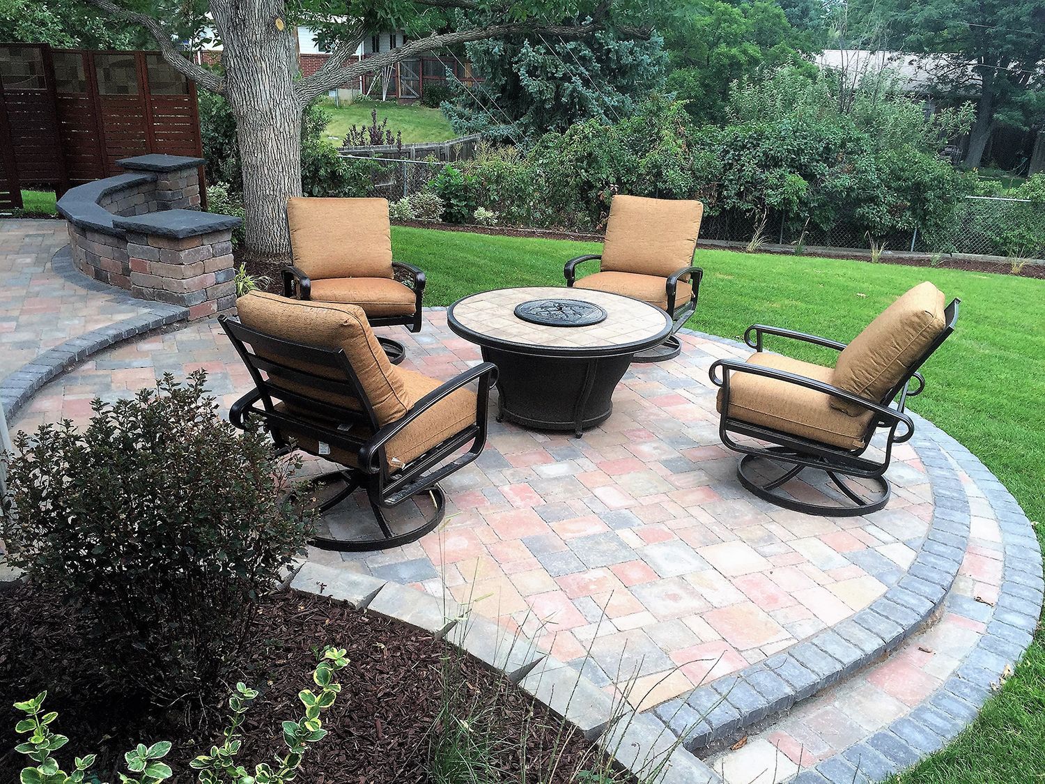 A curved stone patio with a fire pit and four cushioned metal chairs, set in a backyard near a garden and low stone wall.