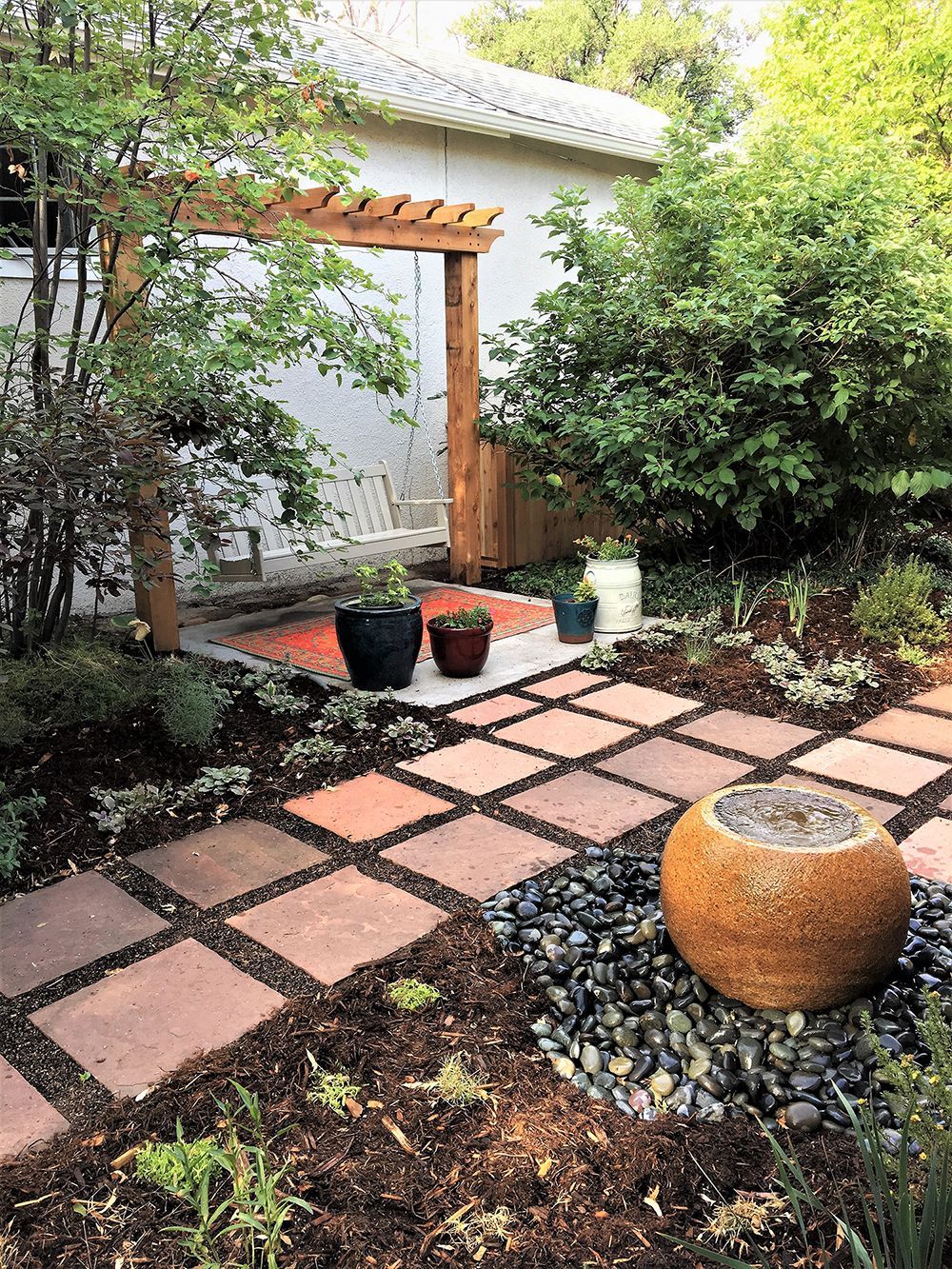 A garden path of square terracotta pavers leads to a wooden arbor, accented by a round stone fountain and landscaping.