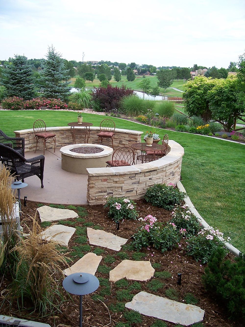 A curved stone wall encloses a patio with a fire pit and metal chairs, set in a backyard garden with stone pavers.