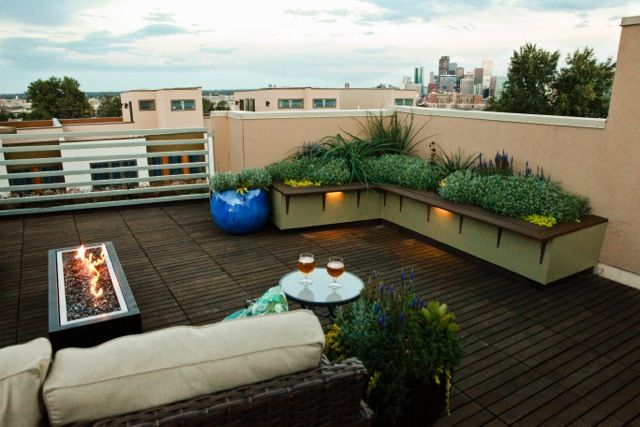 Rooftop patio with built-in seating, plants, fire pit, table, and city view at dusk.