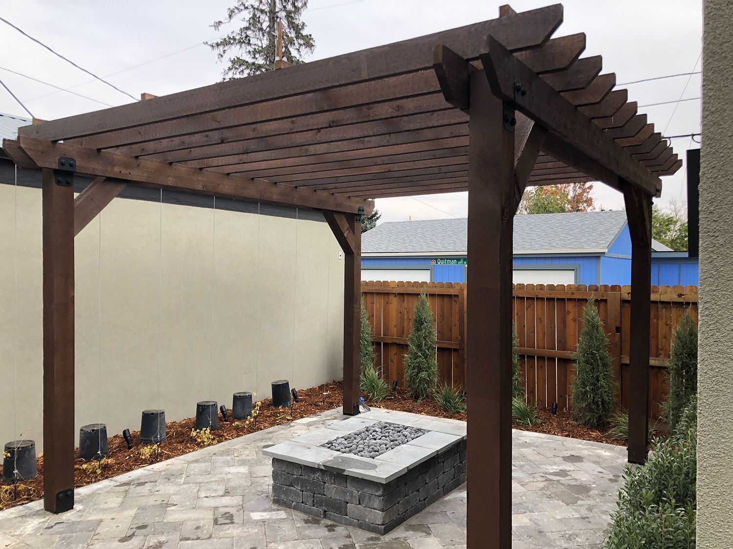 A dark wood pergola covers a stone fire pit on a paved patio in a residential backyard.