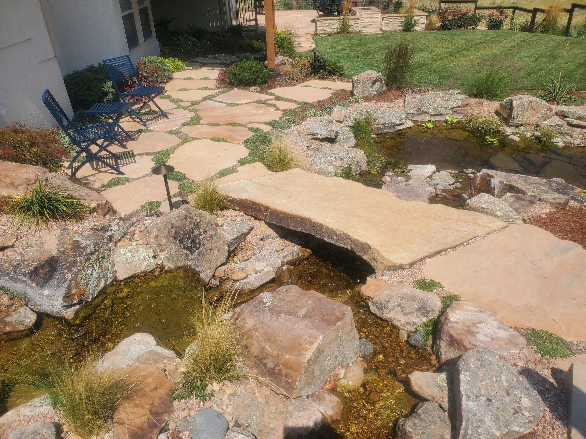 A backyard patio with flagstone paving, a small stone bridge, and a rocky pond surrounded by grass and landscaping.