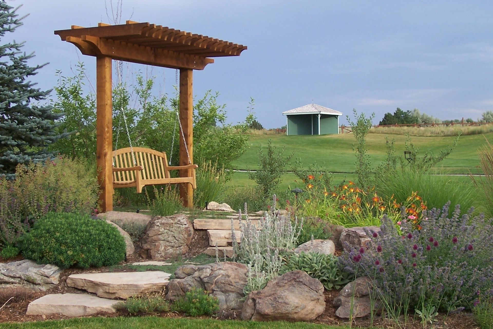 Wooden swing under a pergola in a garden, with a small green gazebo in the distance under a cloudy sky.