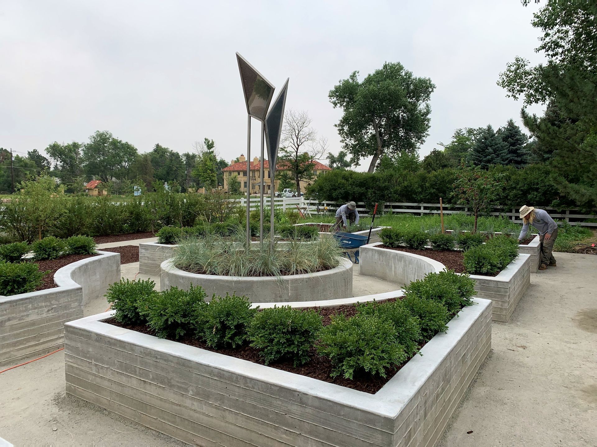 Garden beds with concrete borders, a sculpture, and two people tending plants outdoors.