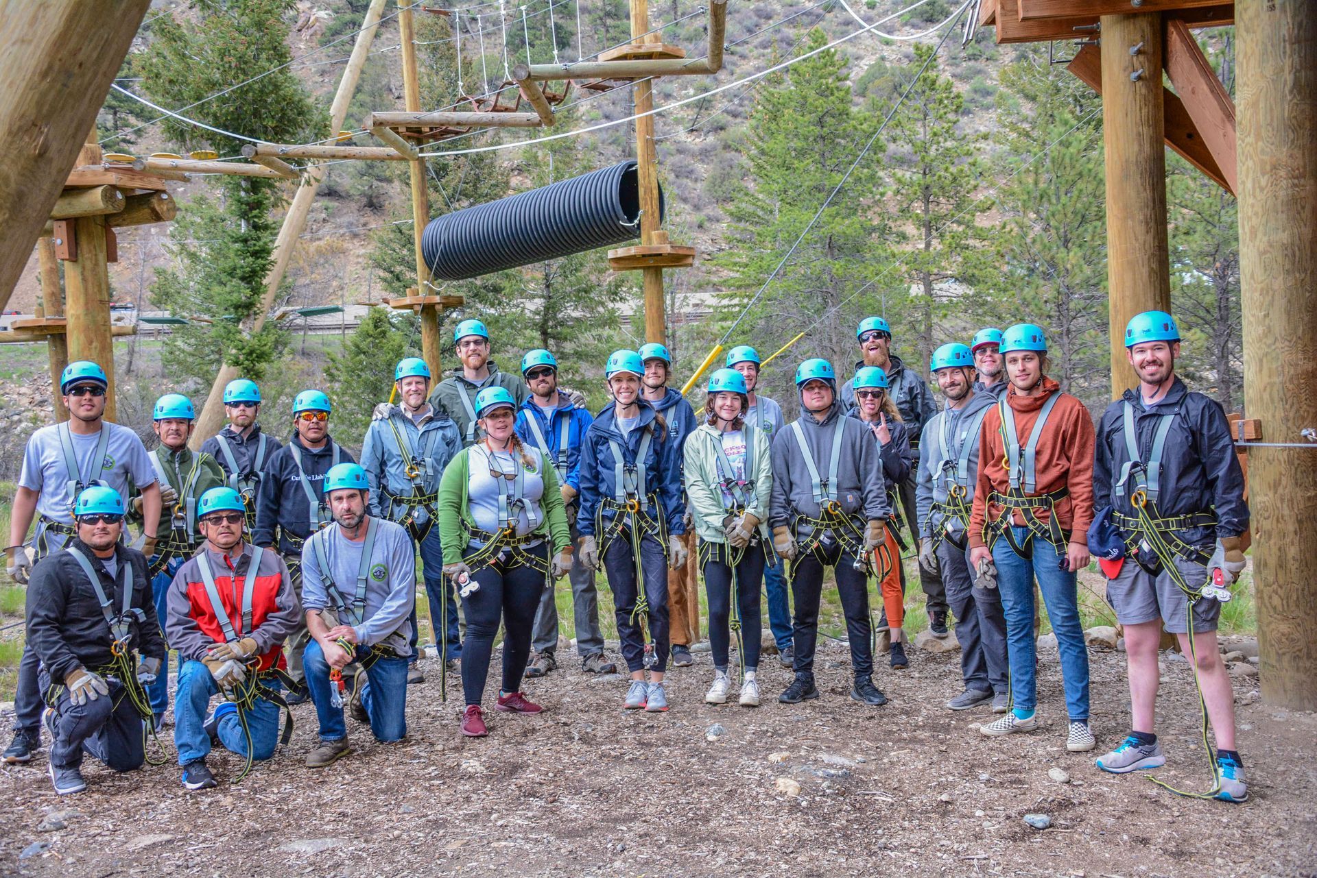 Group of people in harnesses and helmets on a ropes course. They are standing in front of the course, smiling.