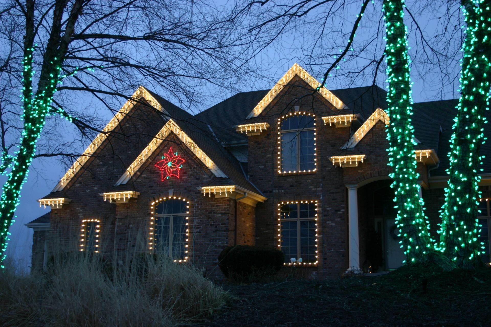 House decorated with Christmas lights, including rooflines and a star, with green lights on trees.