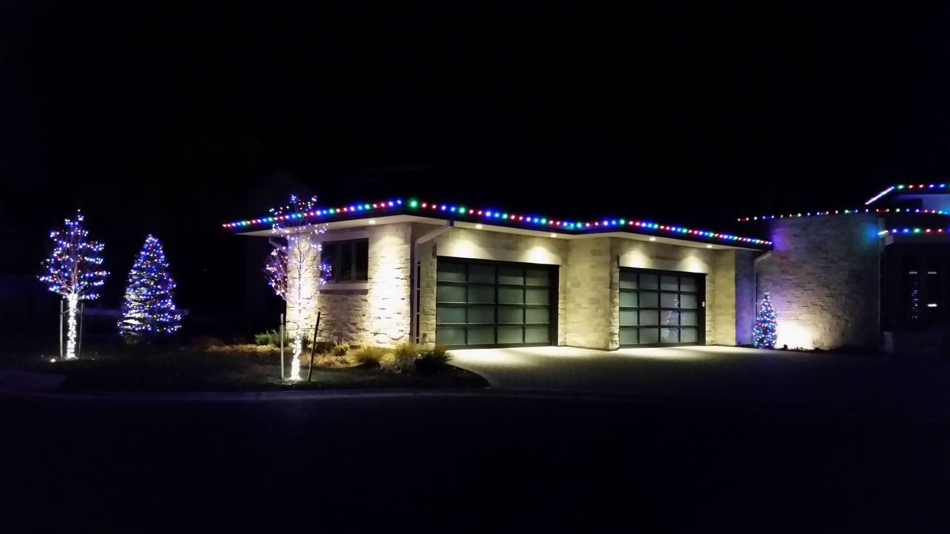 A house lit with colorful Christmas lights at night, trees decorated, garage doors illuminated.