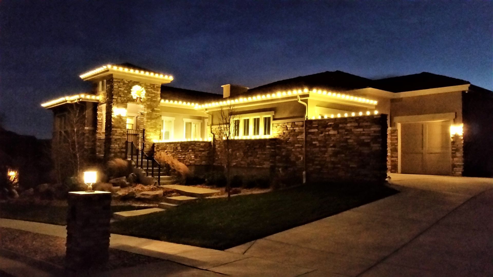 House exterior at night, decorated with warm white lights; stone facade and garage.