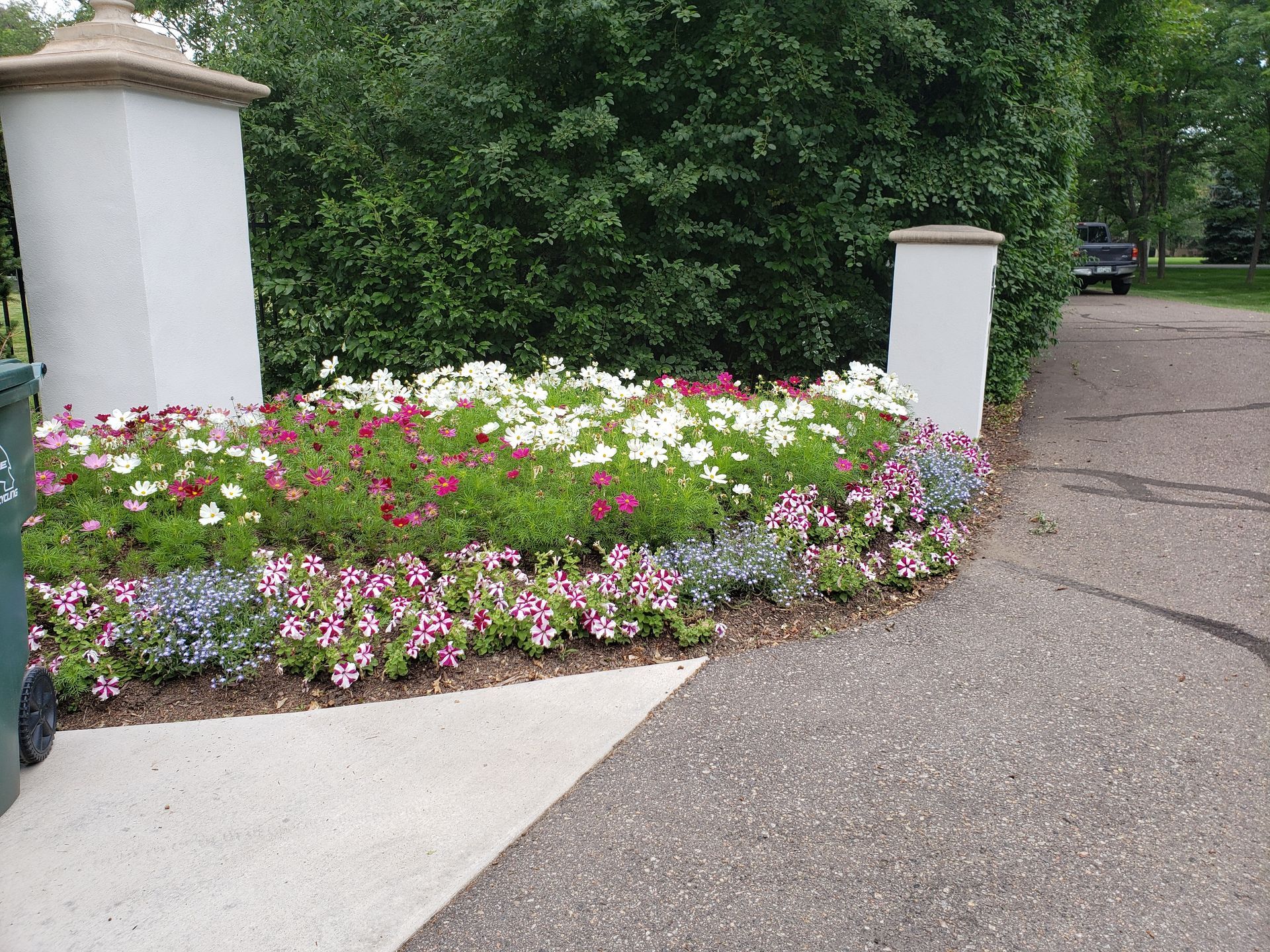 Flowering garden bed at a driveway entrance with white, pink, and purple blooms, between two white pillars.
