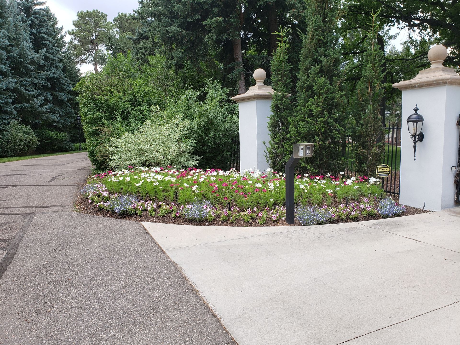 Flower bed in front of white pillars with gate; driveway to the left, trees in background.