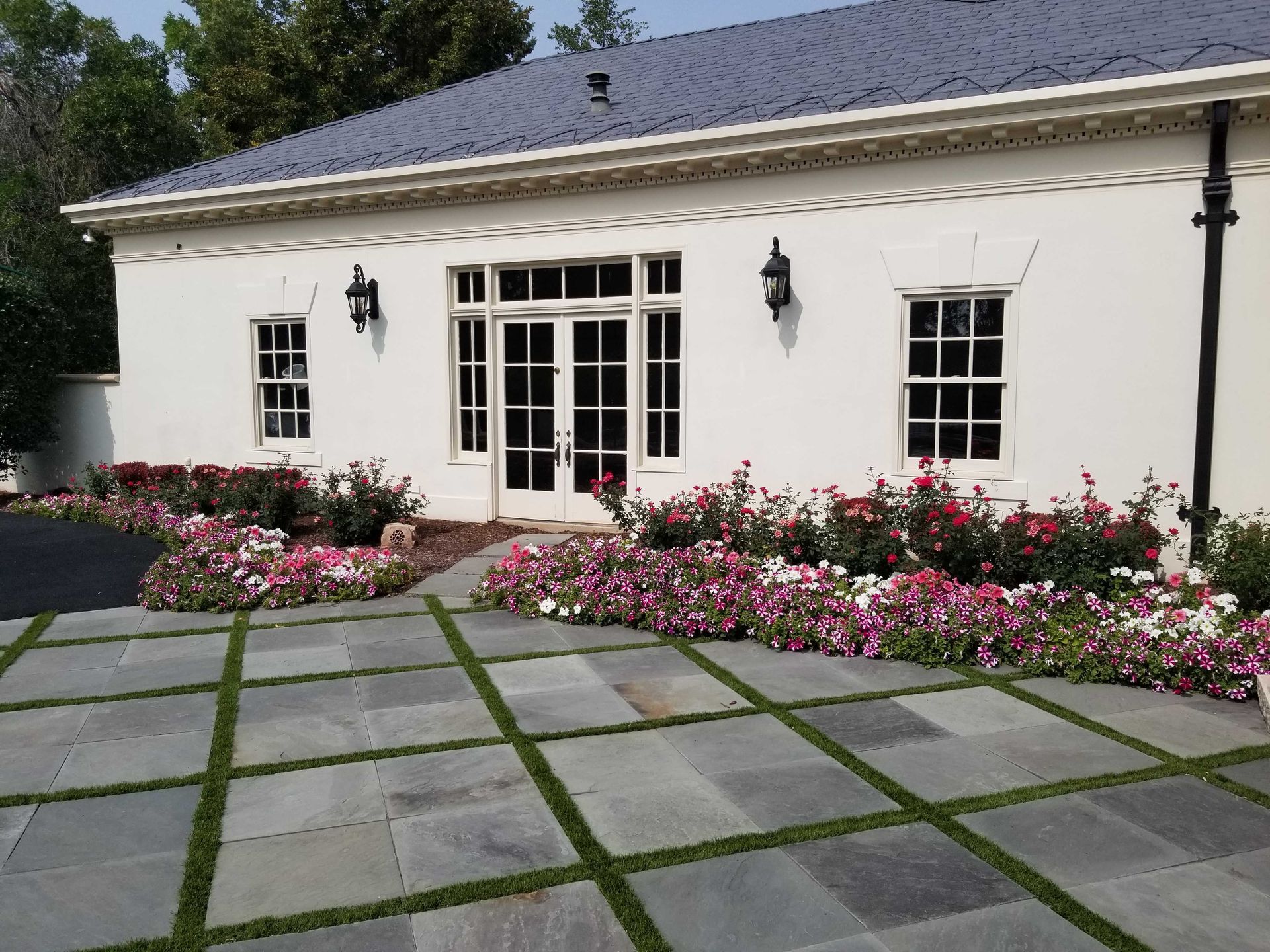 White building with black roof, flanked by pink and white flowers. Paved patio with green grass squares.